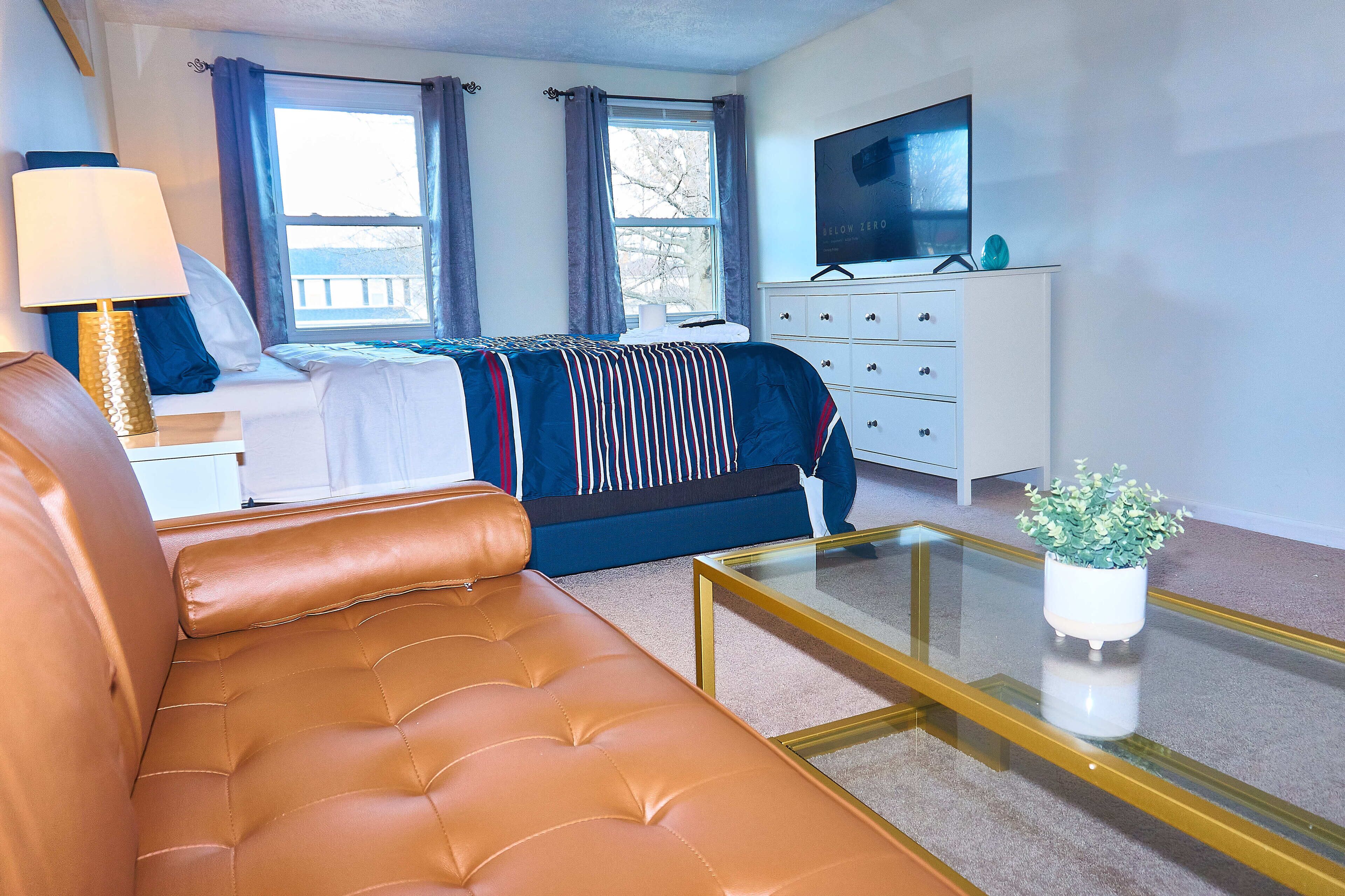 Quarto de hotel moderno com uma cama, um sofá, uma televisão, uma cômoda e uma pequena planta sobre uma mesa de vidro.