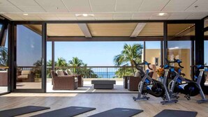 Un gimnasio con bicicletas estáticas, colchonetas negras, mobiliario de mimbre y vistas al océano.