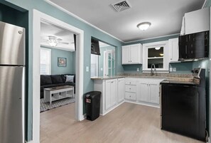 Kitchen features a stainless steel refrigerator, black oven, and white ceiling fan, adjacent to a living area with a gray couch and coffee table.