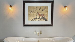 A freestanding bathtub in a room with wooden flooring, a framed painting of birds and a fox on the wall, and two wall-mounted lights. - Stylish apartment above popular traditional village pub! (Saint Brides Major)