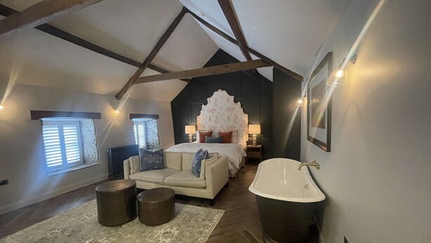 A bedroom with a large bed, a sitting area with a sofa, a bathtub, and a window with blinds. - Stylish apartment above popular traditional village pub! (Saint Brides Major)