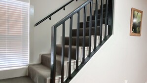 Uma escadaria com corrimão preto, degraus carpetados e parede branca. Há três quadros na parede.
