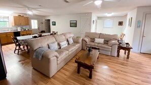 Uma sala de estar espaçosa com um amplo sofá seccional bege, uma mesa de centro de madeira e um ventilador de teto. Há uma área de cozinha com armários de madeira e uma mesa de jantar.