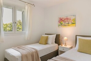 A bedroom with two beds, a window with curtains, a nightstand with a lamp, and a painting on the wall.