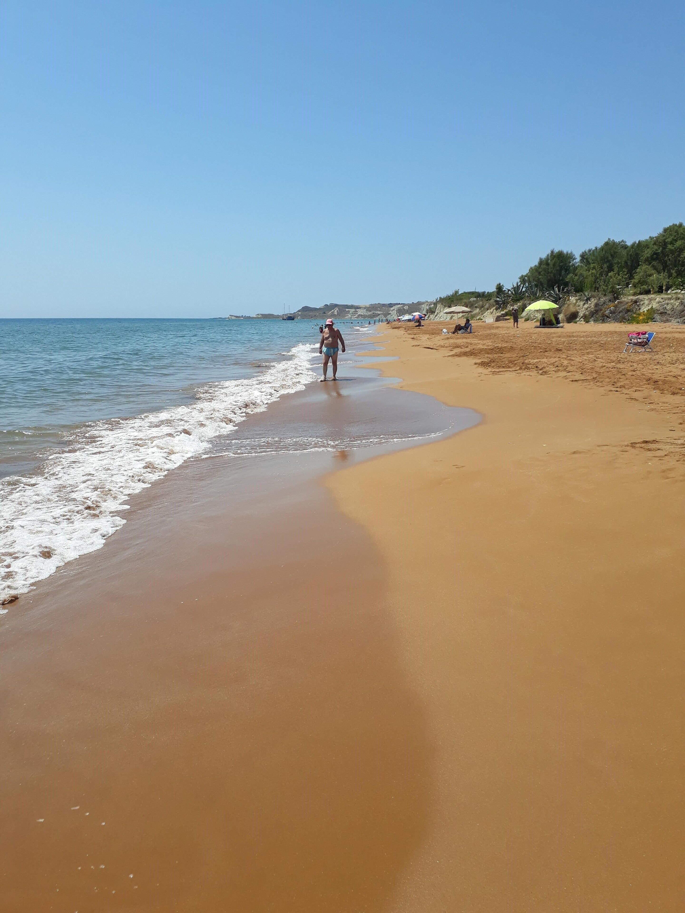 Ein Strand mit sandigem Ufer, einigen Sonnenschirmen und einer Person, die am Wasser entlanggeht.