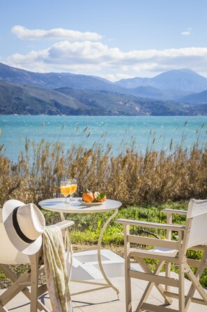 A table with two glasses of orange juice, a plate with oranges, and a hat on a chair, overlooking a turquoise lake with mountains in the background.