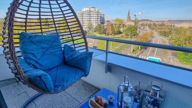 A balcony with a hanging chair, a small table with a croissant and coffee, and a view of buildings and greenery. - Exceptional apartment in Strasbourg (Strasbourg)