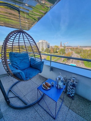 A balcony with a hanging chair, a small table with a croissant and coffee, and a view of buildings and greenery.
