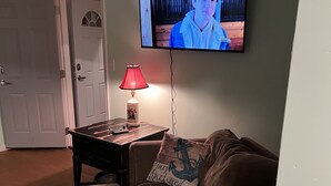 Una sala de estar con un televisor montado en la pared, una mesita con una lámpara y un sofá marrón con un cojín de diseño náutico.