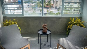 A room with two chairs facing a small table with a decorative item, a large window with a view of buildings and greenery, and a concrete wall. - 3 BR Residence w. Private Terrace + Rooftop (San Juan)