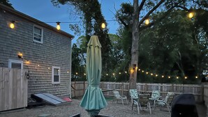 Un jardin avec une table et des chaises sous une pergola, un barbecue et des guirlandes lumineuses.