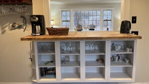 Un comptoir de cuisine avec une machine à café, un panier et des verres à vin. Des étagères situées en dessous contiennent divers ustensiles de cuisine, et une fenêtre offre une vue sur les arbres.