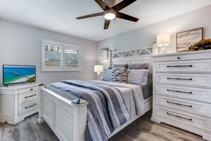 A bedroom with a bed, a TV, a ceiling fan, a window with blinds, and a dresser.