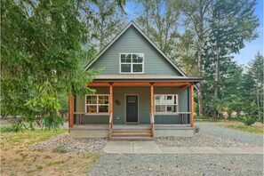 Charming two-story house with a gabled roof and inviting front porch, surrounded by lush trees and a gravel driveway.