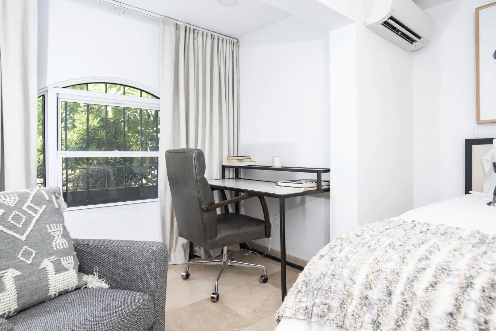 A modern hotel room with a bed, a desk, and a chair. There is a window with curtains, an air conditioning unit, and a view of greenery outside.