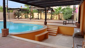 Una piscina con bordo curvo, una zona patio coperta con poltrona relax e un angolo barbecue.