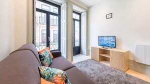 Een moderne woonkamer met een bruine bank, kleurrijke kussens, een televisie op een houten dressoir en grote ramen met uitzicht op gebouwen.