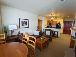 A living room with a sofa, chairs, a coffee table, and a dining table. There is a kitchen in the background with a microwave, stove, and cabinets.