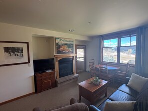 A living room with a fireplace, a television, a dining table with chairs, and a large window with a view.