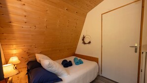 A cozy bedroom with a wooden ceiling, a bed with a white cover, two rolled towels, a blue pillow, a wall-mounted light, and a door with a simple handle. - Nouveau chalet romantique à la montagne avec jacuzzi et sauna privatif (Métabief)