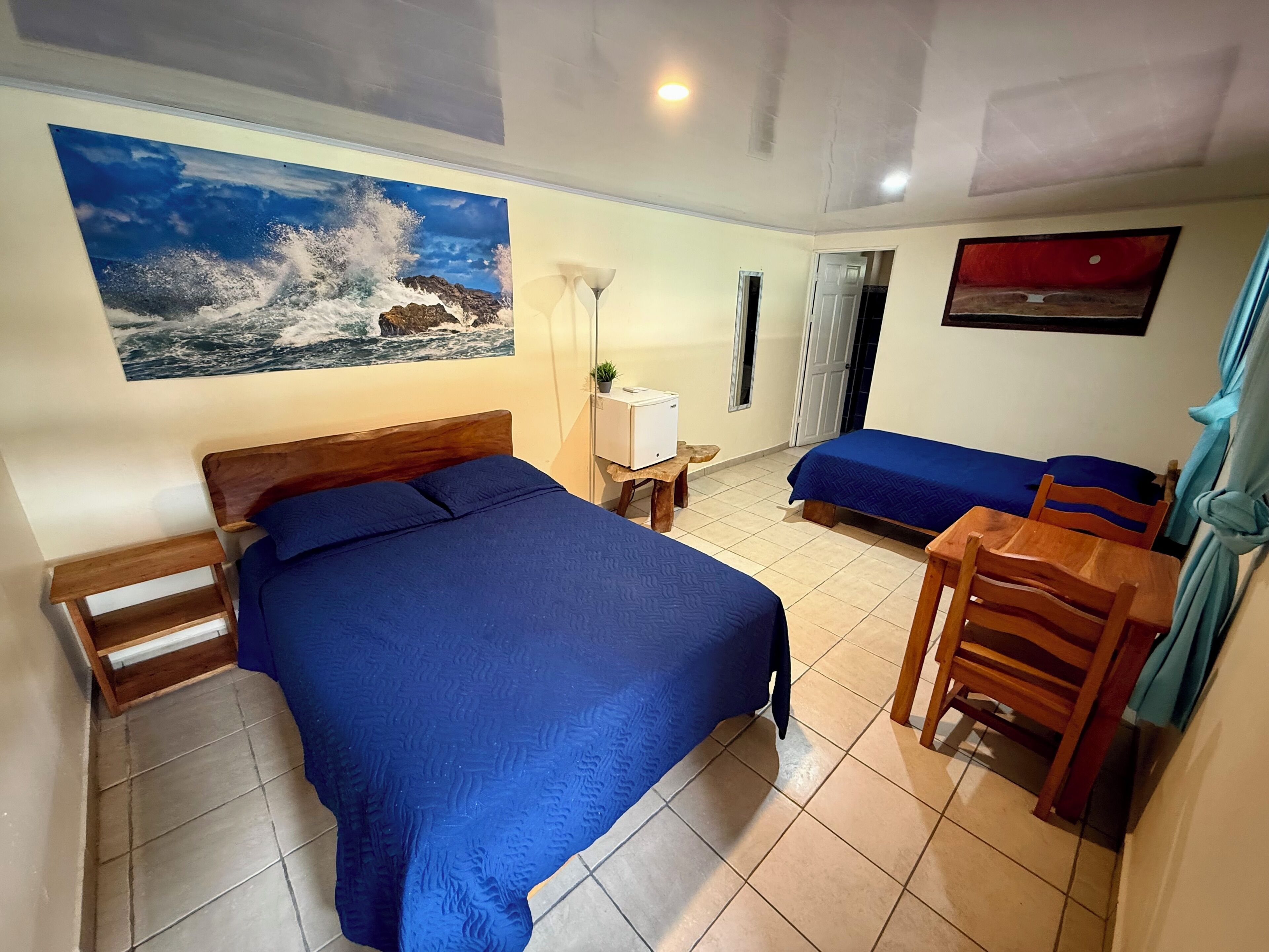 A hotel room with a large bed, a smaller bed, a desk with a chair, a TV, and a painting of a wave crashing against rocks.
