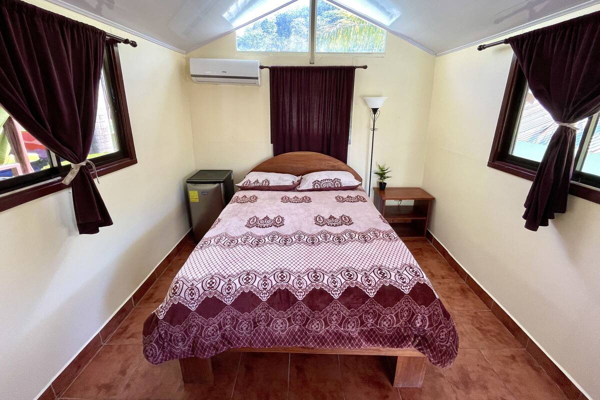 A single-bed room with a patterned bedspread, a small bedside table with a lamp, a window with curtains, and an air conditioning unit on the wall.