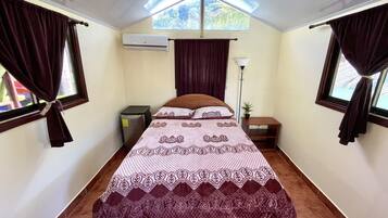 Une chambre avec un lit simple, une couvre-lit à motifs, une petite table de chevet avec une lampe, une fenêtre avec des rideaux et un appareil de climatisation fixé au mur.