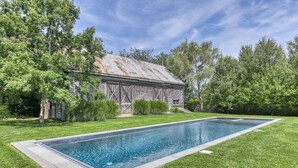 Una piscina rectangular con borde de piedra, rodeada de un césped cuidado y un granero rústico de madera con techo metálico.