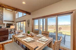 A dining area with a wooden table set for four, large windows offering a view of the outdoors, and a stone fireplace. - Mimosa Fields: Designer Kitchen w/ Outdoor Oasis (Fruita)