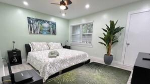 Quarto com cama, mesa de cabeceira, escrivaninha, planta em vaso e janela com persianas.