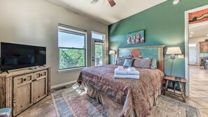 A bedroom with a large bed, a television on a wooden cabinet, and a view of the outdoors through a window. - Scenic Ruidoso Home w/ Large Deck + Hot Tub! (Ruidoso)
