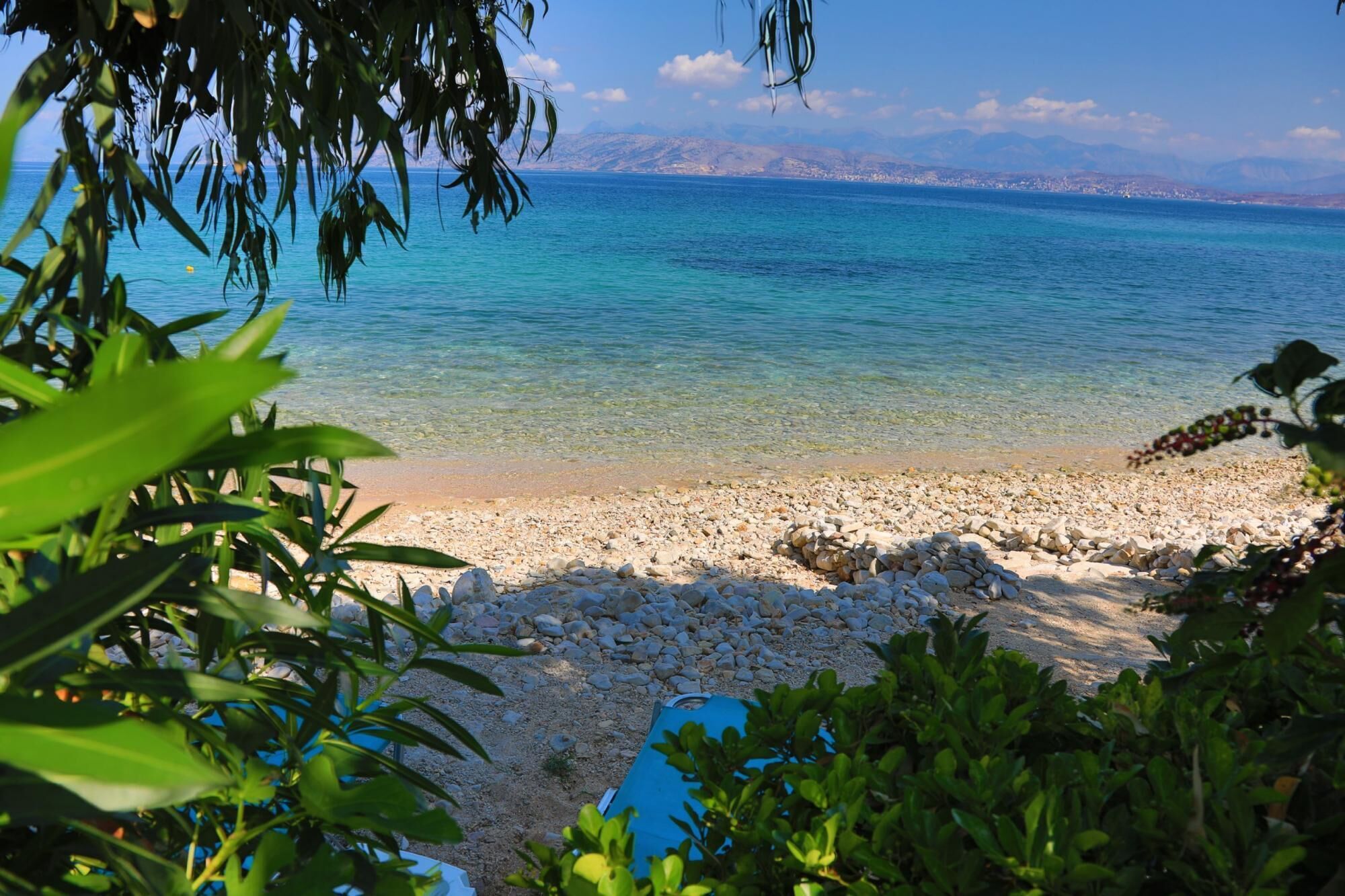 A beach with clear turquoise water, pebbly shore, and distant mountains.