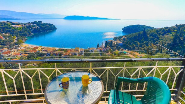 A balcony with a glass table and chairs overlooking a coastal landscape with a clear view of the sea and distant hills. - The Old Office in Kalami (Corfu)