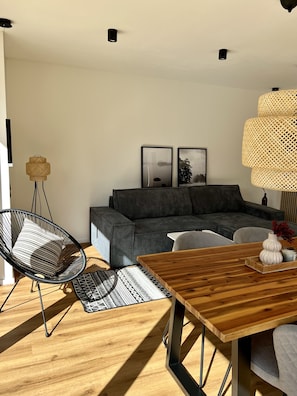 Ett modernt vardagsrum med ett soffbord i trä, en svart stol med metallram, en grå soffa och en golvlampa med flätad skärm.