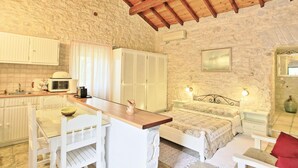 A rustic room with a stone wall, wooden ceiling beams, a bed with a floral bedspread, a small table with chairs, a kitchen area with a microwave, and a sink. - Korinas Cottage in Kentroma (Corfu)