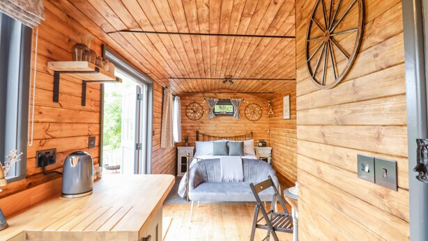 Een compact houten hutje met een keukenhoekje, een bed met hoofdeinde, en een grote houten wieldecoratie aan de muur.