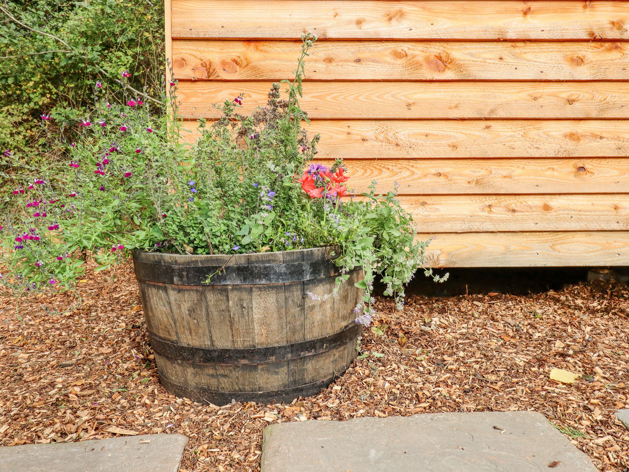 A wooden barrel planter filled with various plants and flowers, placed on mulch, next to a wooden fence.