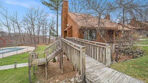 Eine Holzterrasse führt zu einem Haus mit Schornstein, umgeben von Bäumen und einem eingezäunten Bereich.