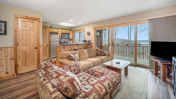 Ein gemütliches Wohnzimmer mit einem großen Sofa, einem Couchtisch und einem Flachbildfernseher. Es gibt eine Küchenzeile mit Holzausstattung und einem Kühlschrank. Das Zimmer verfügt über eine große Glasschiebetür, die auf einen Balkon mit Blick auf Bäume führt.
