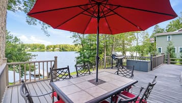 A deck with a table and chairs, overlooking a lake with a view of trees and a house in the background.