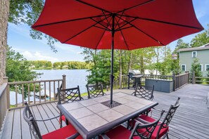 A deck with a table and chairs, overlooking a lake with a view of trees and a house in the background.