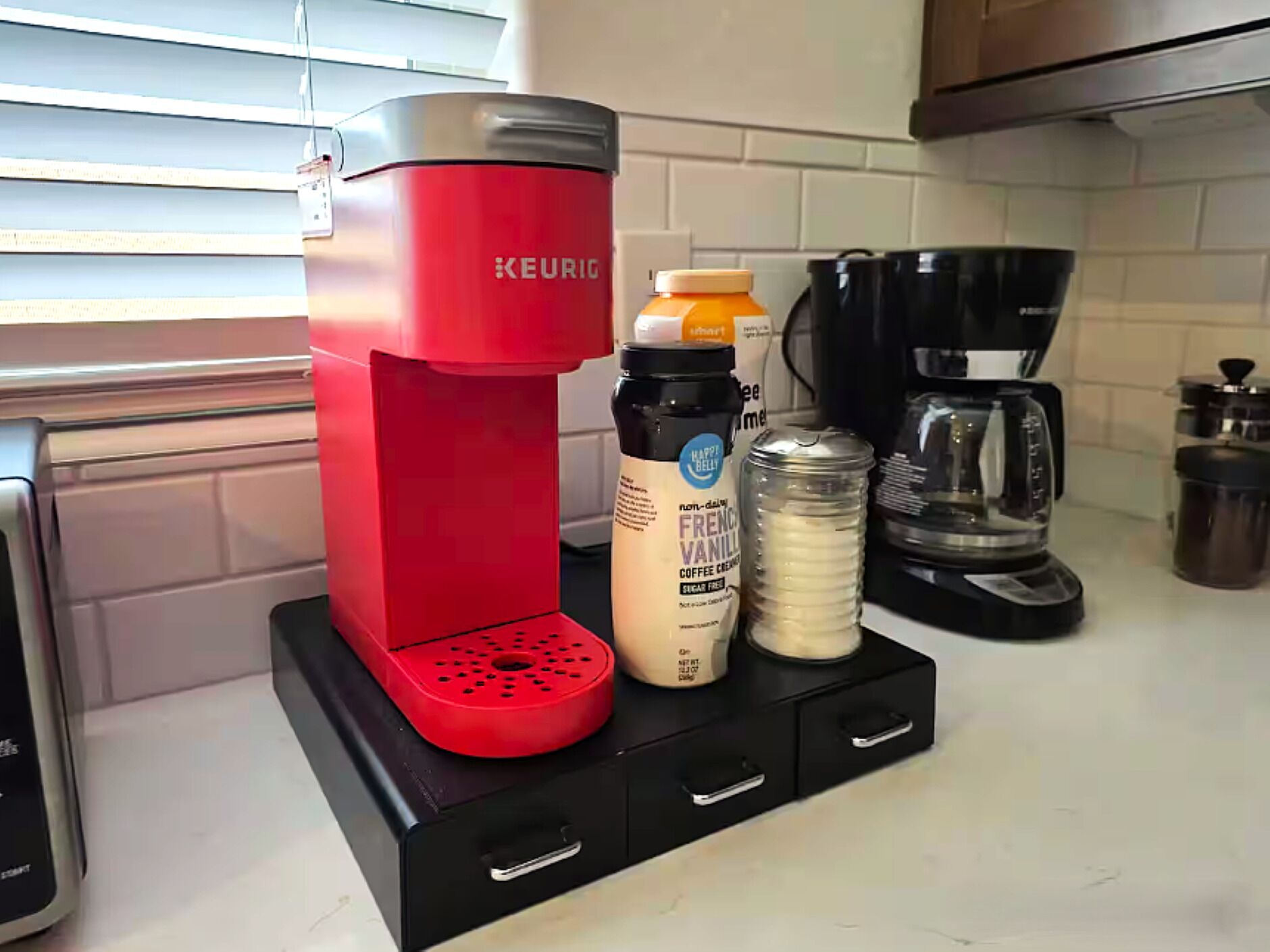A red Keurig coffee maker on a black base with a coffee pod, a bottle of French Vanilla coffee creamer, and a container of creamer on a kitchen counter.