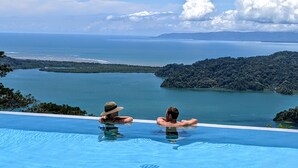 Un couple se détend dans une piscine à débordement, avec vue sur une vaste étendue d’eau et une île boisée.
