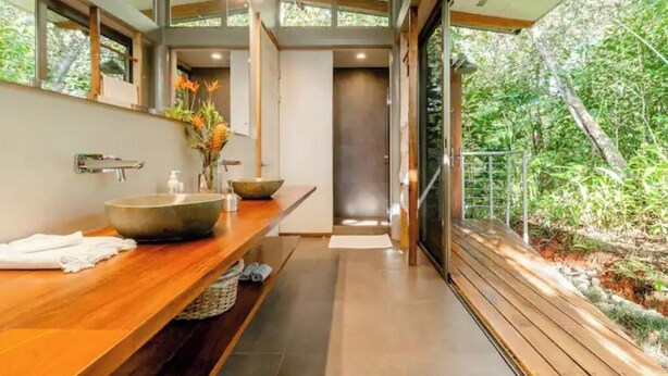 Une salle de bain moderne avec un grand meuble-lavabo en bois, un lavabo en forme de bol et un miroir. On peut admirer la verdure à travers la fenêtre.