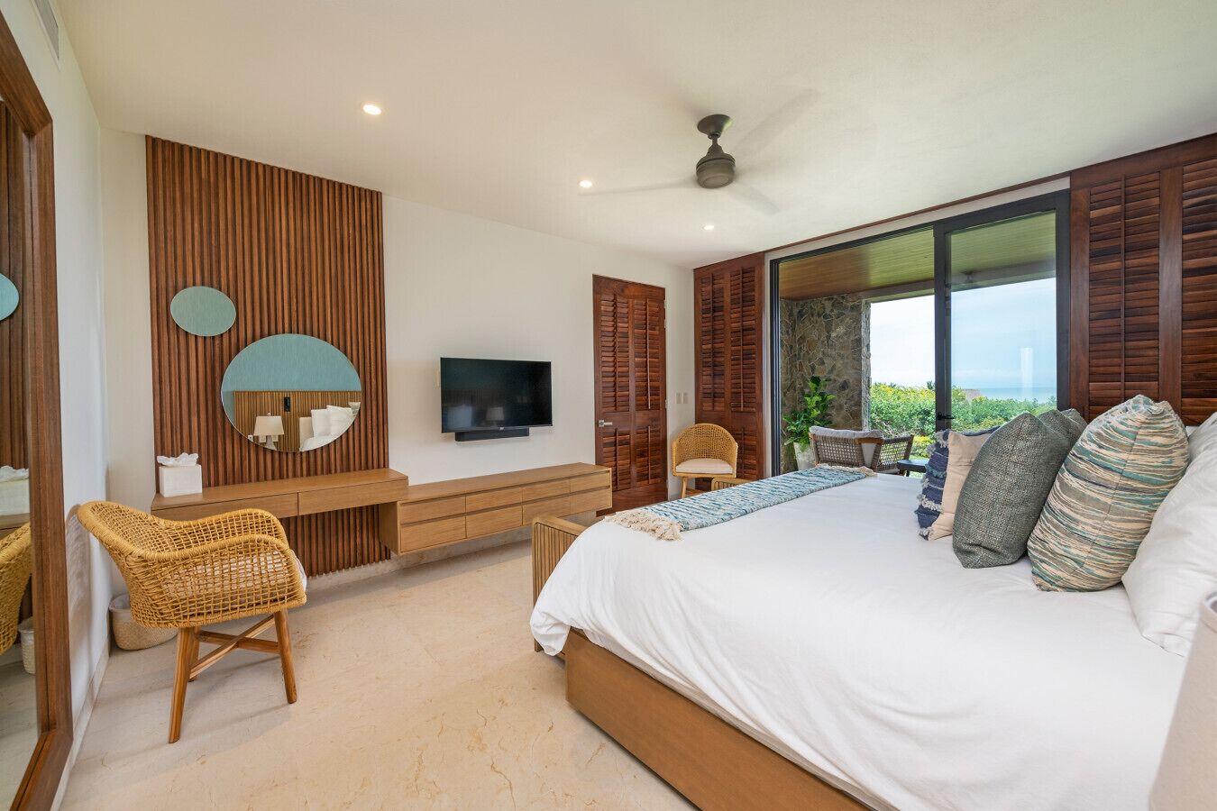 A bedroom with a large bed, a TV, a wooden vanity, and a view of the outdoors.