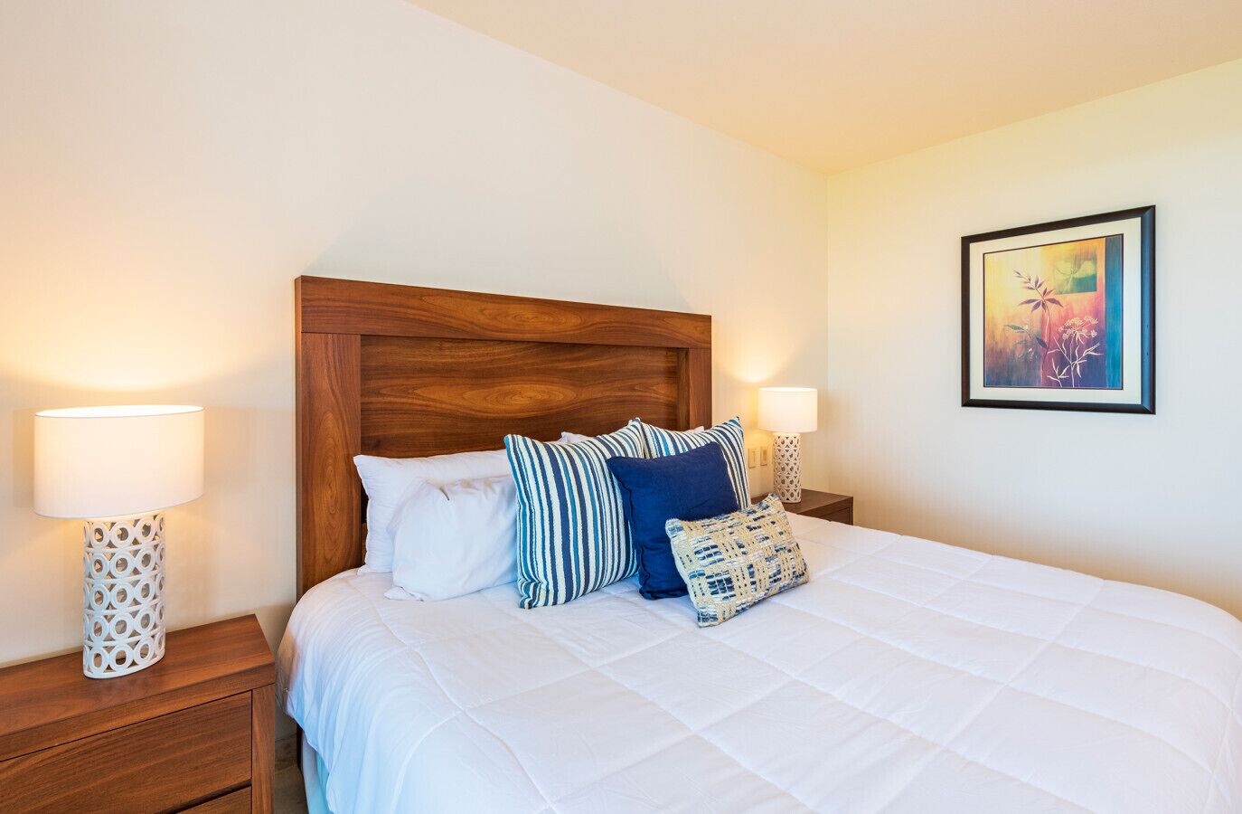 Een net opgemaakt bed met witte beddengoed en meerdere kussens, twee bedlampen en een lijst met een kunstwerk aan de muur.