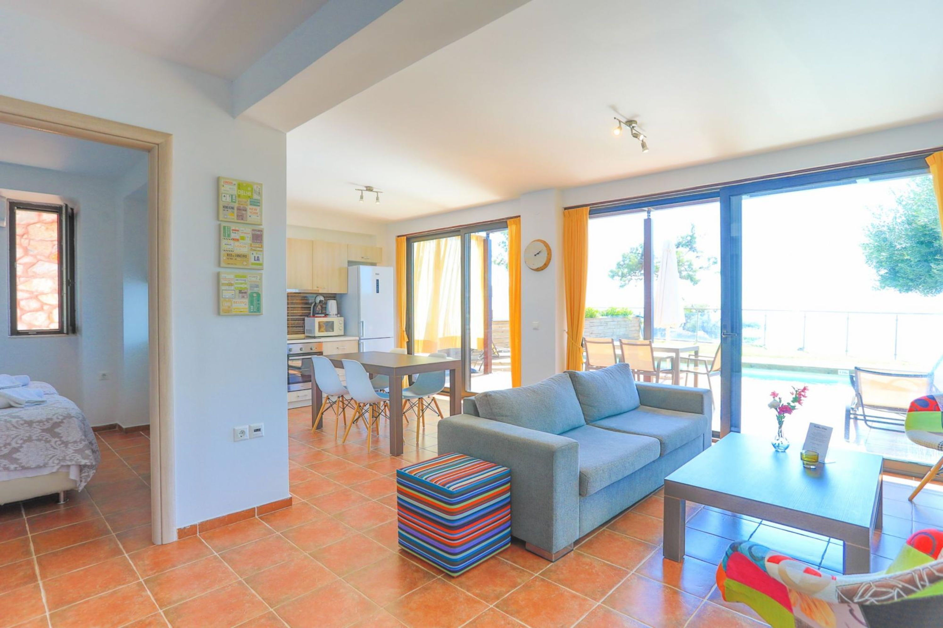 Un salon moderne avec un canapé, un pouf et un coin repas. On aperçoit une cuisine à l’arrière-plan, avec une table et des chaises.