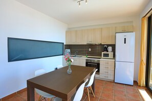 A modern kitchen with a dining area, featuring a large flat-screen TV, a wooden dining table, white chairs, and a refrigerator. - Kalma Villa Ena in Moudania (Nea Propontida)