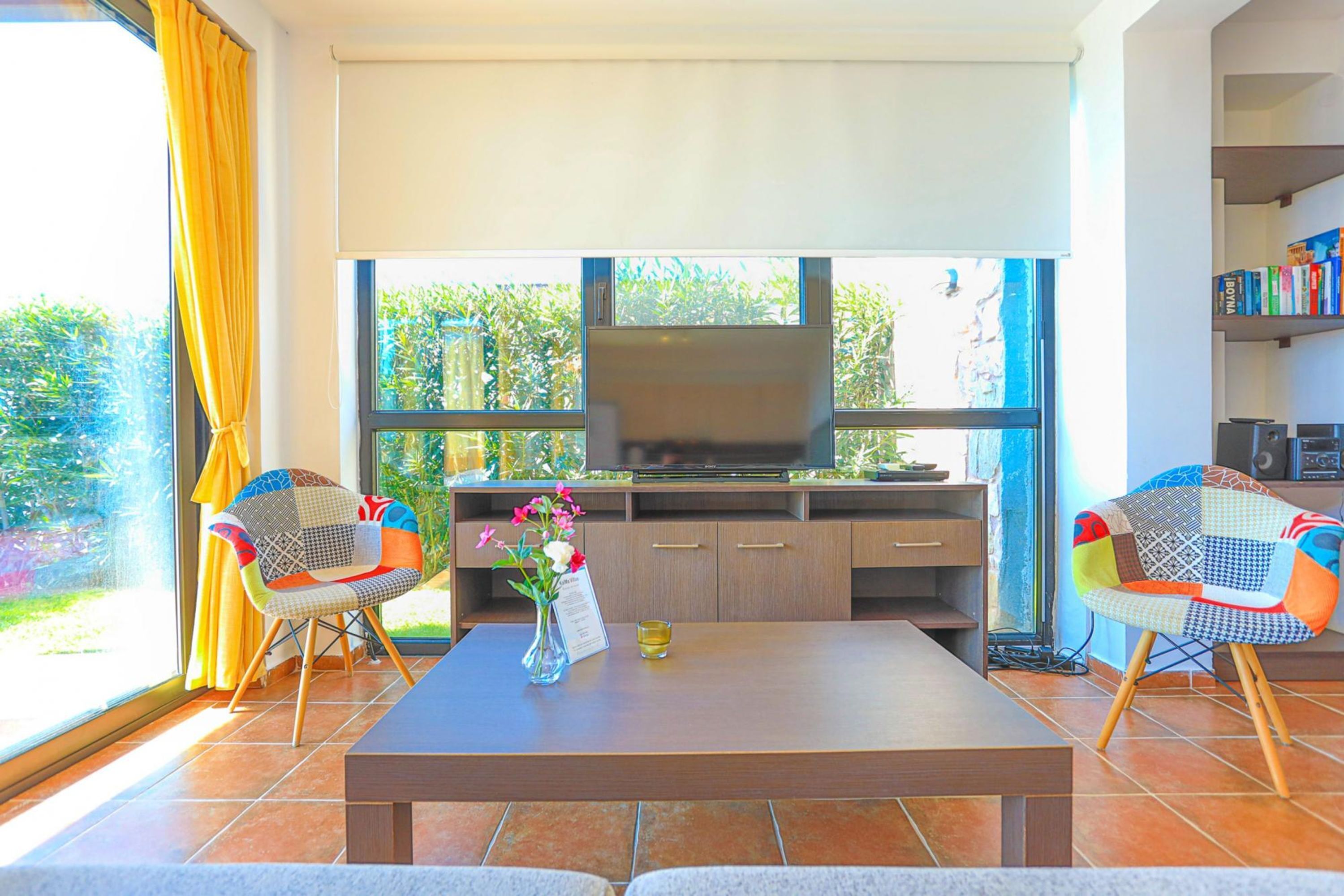 A modern living room with a large TV, two colorful chairs, a coffee table, and a bookshelf.