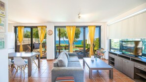 A modern living room with a sofa, dining table, chairs, and a TV. Large windows offer a view of the sea.